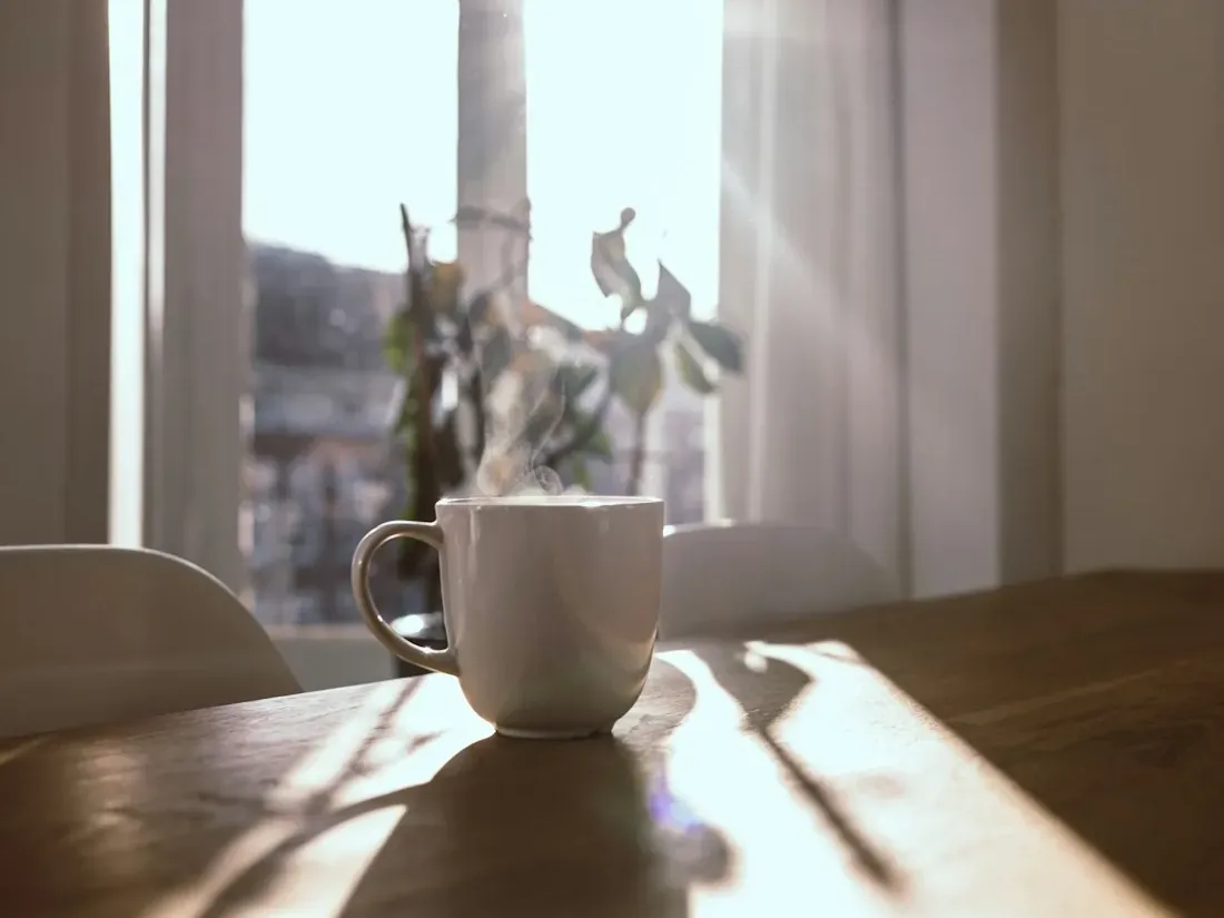 Taza blanca sobre una mesa de madera junto a una ventana con luz de mañana